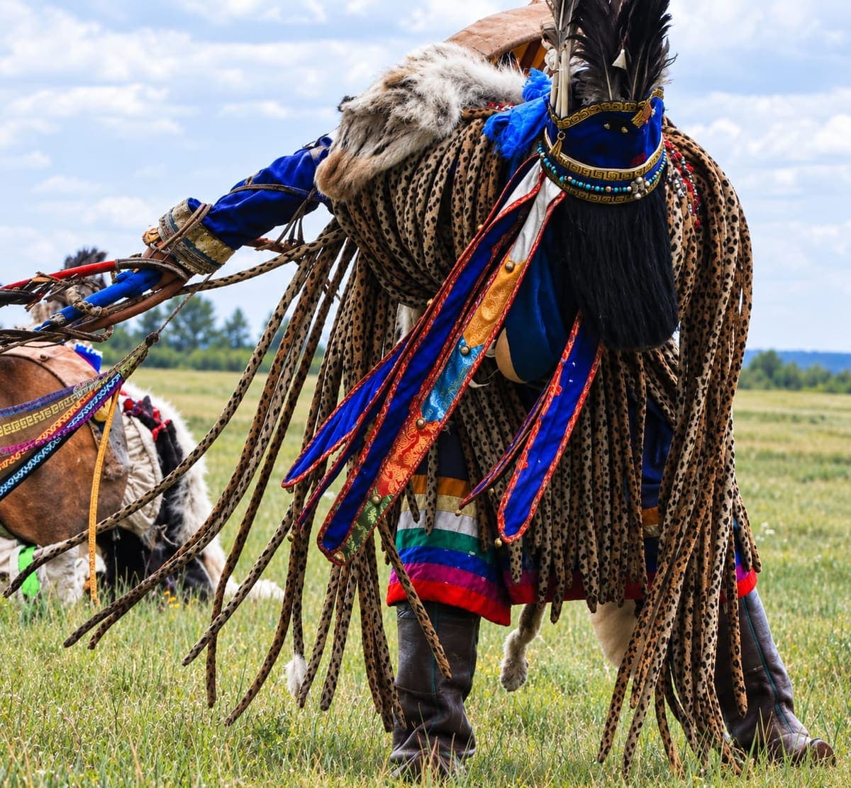 A figure in traditional ceremonial dress with fringed leather and a feathered headdress, walking beside a horse on the open steppe