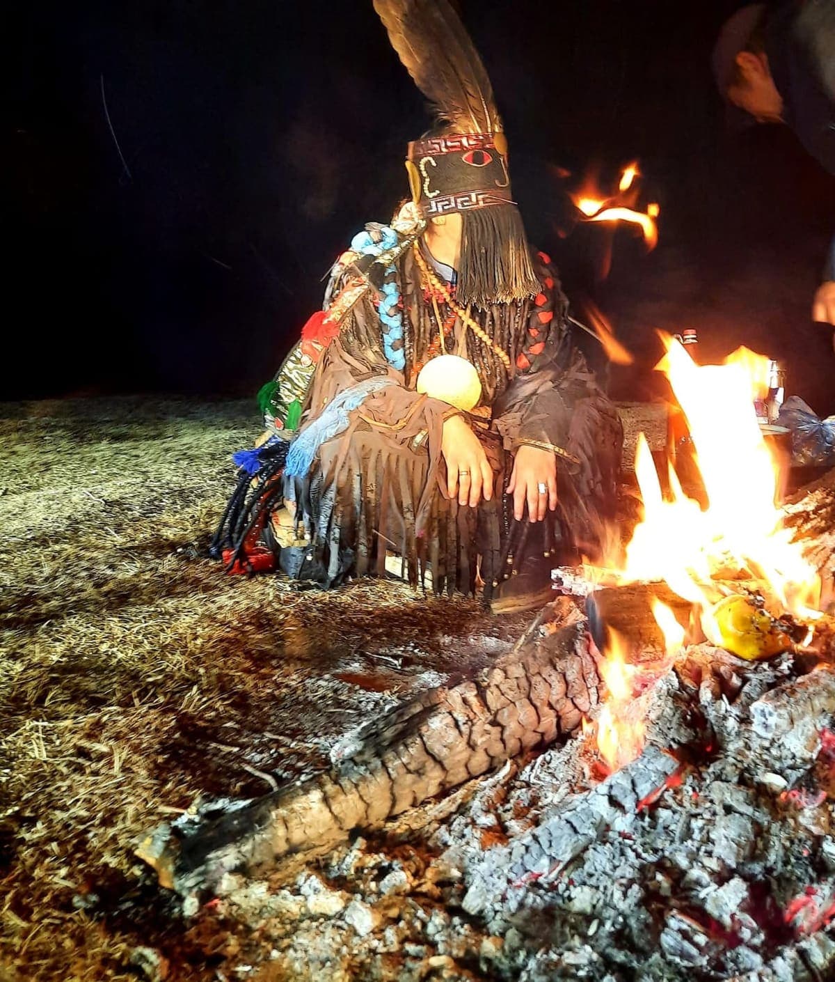 A shaman in ceremonial dress and feathered headdress performing a ritual beside a fire at night